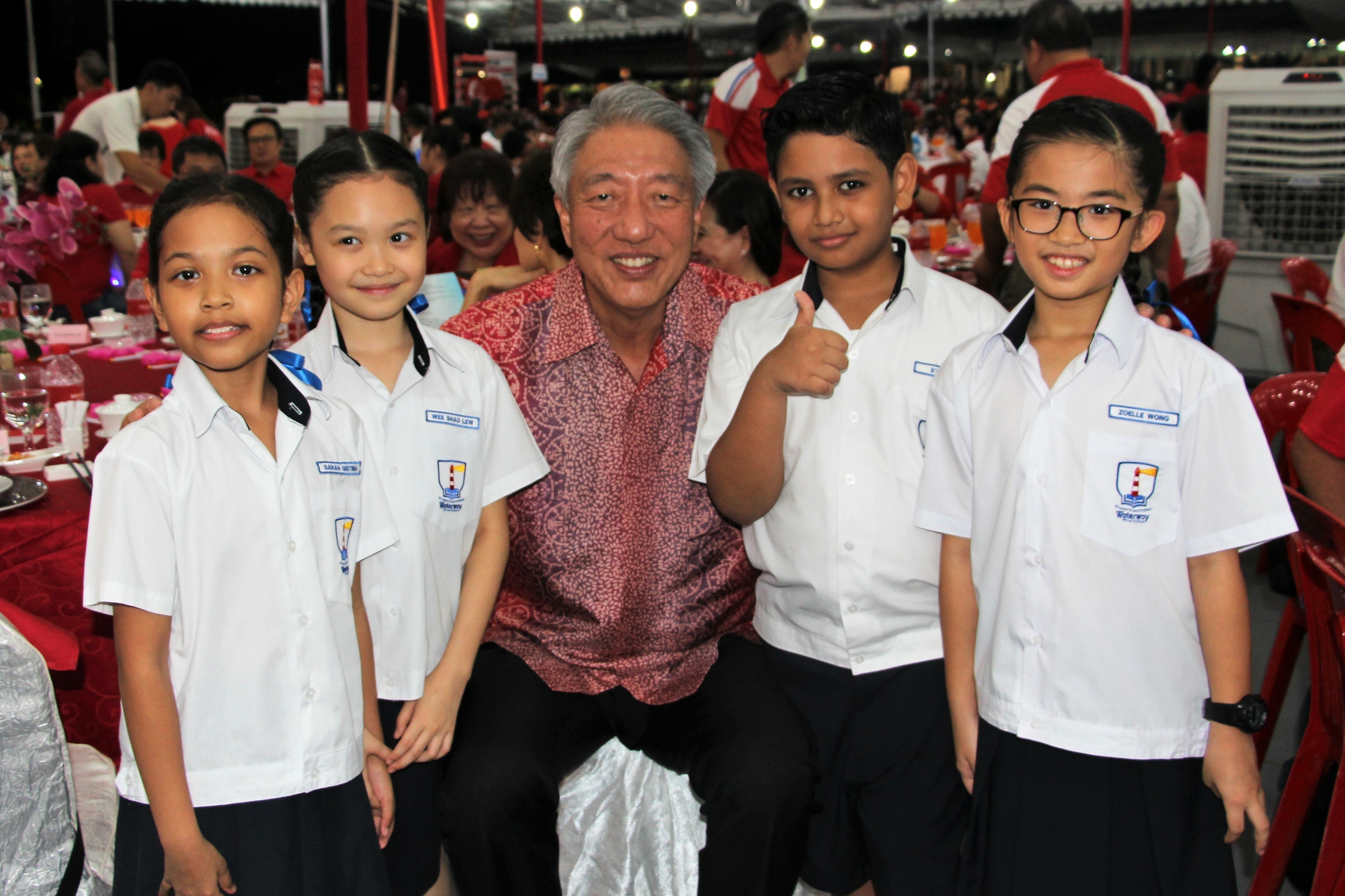 An older man in a red batik shirt gives a thumbs up while posing with four smiling school children in white uniforms.